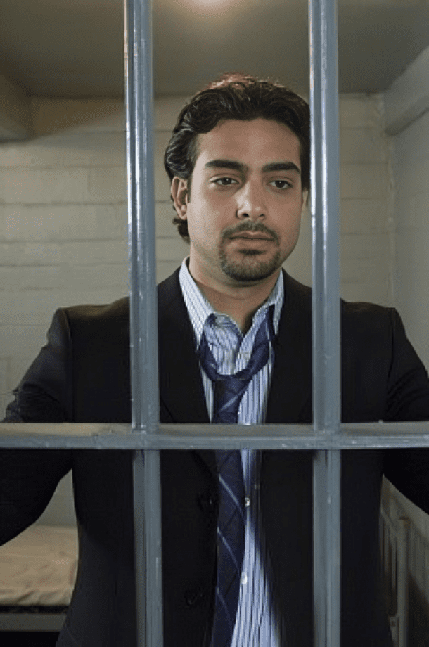 A man in a suit stands behind bars holding onto them, with a somber expression, inside a jail cell.