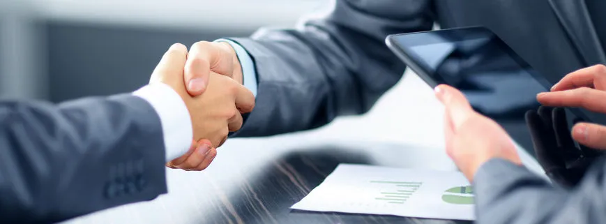 Two businesspeople shaking hands over a desk with documents and a tablet, signifying an agreement or deal.