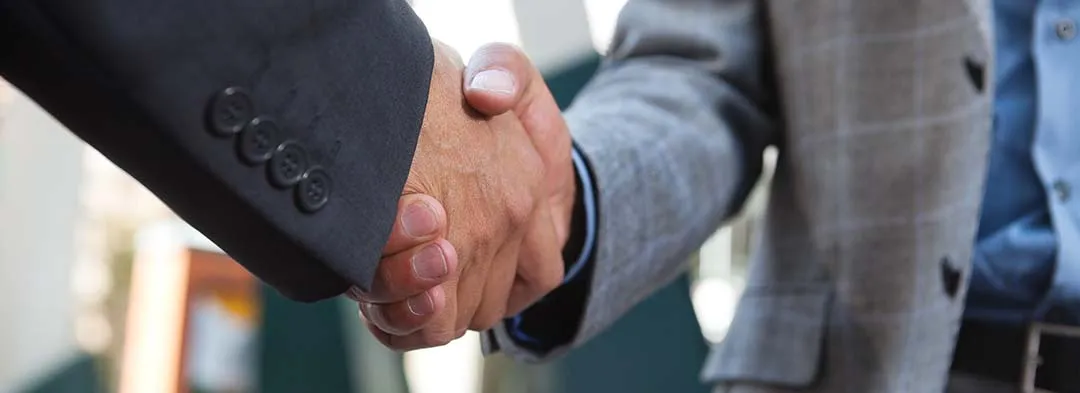 Two people wearing business attire are shaking hands, with one person in a dark suit and the other in a light grey blazer. The background is blurred and indistinct.