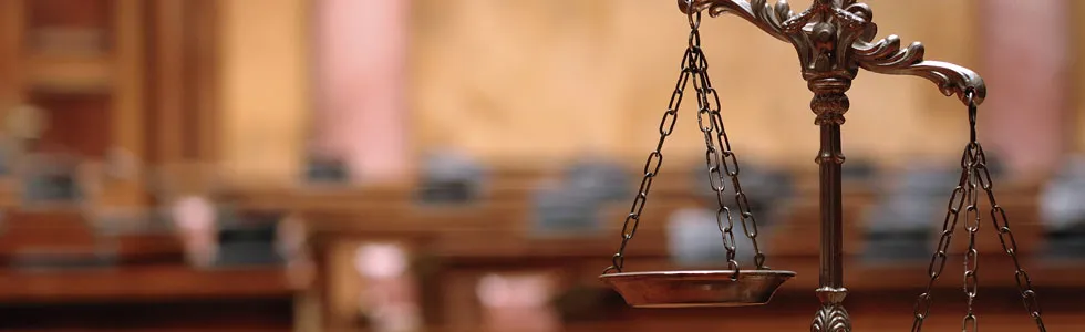 Close-up of a brass balance scale in a courtroom setting, with blurred wooden furnishings and seats in the background.