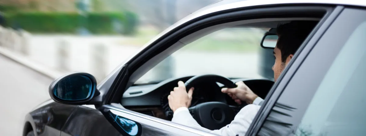 A person wearing a white shirt is driving a black car on a roadway, viewed from the side window. The focus is on the driver's position, holding the steering wheel, with a blurred background visible.