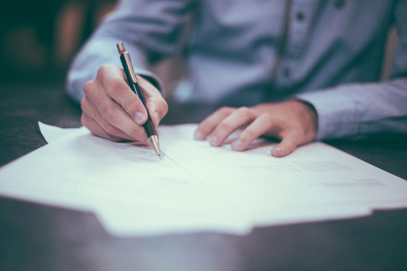 A person in a long-sleeve shirt is writing on papers with a pen while sitting at a desk.