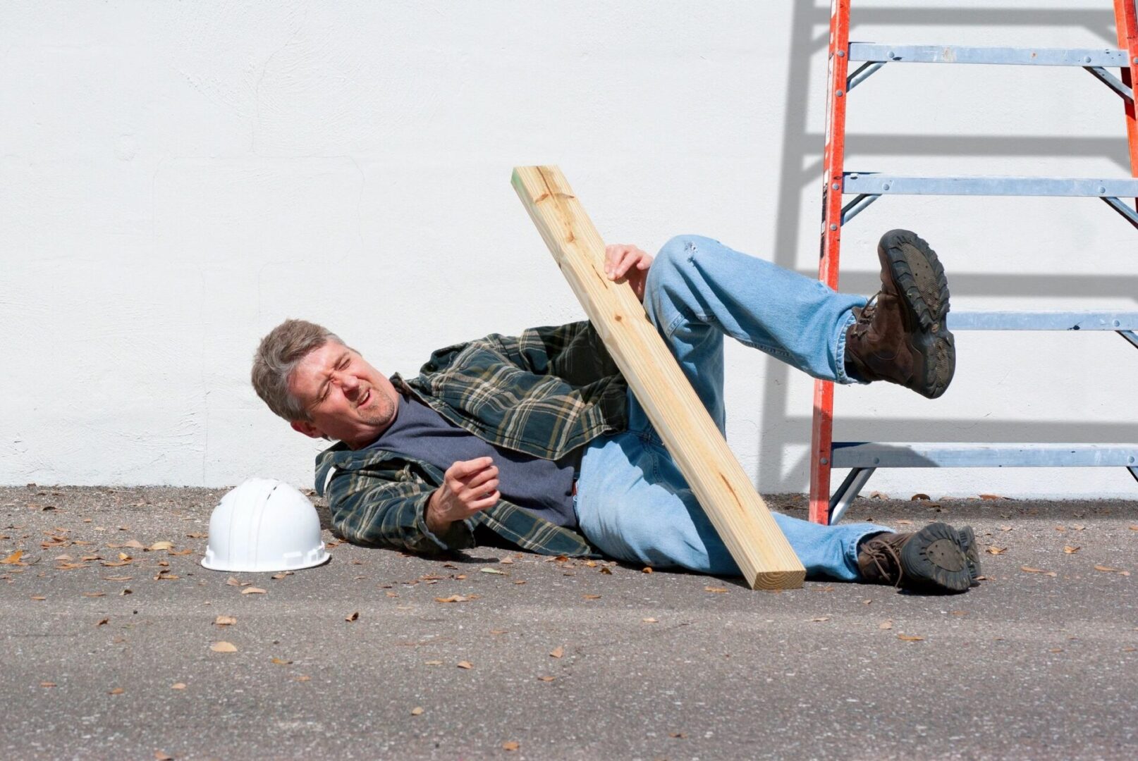 A man wearing a plaid shirt and jeans falls to the ground while holding a wooden board next to a ladder and a white hard hat.