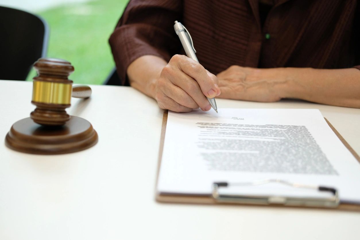 A person signs a document on a clipboard beside a judge's gavel on a white table.