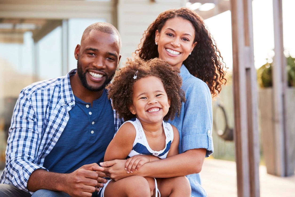 A smiling family of three sits together outdoors. The man and woman are seated closely with a young girl sitting in front of them. All three are wearing casual clothes.
