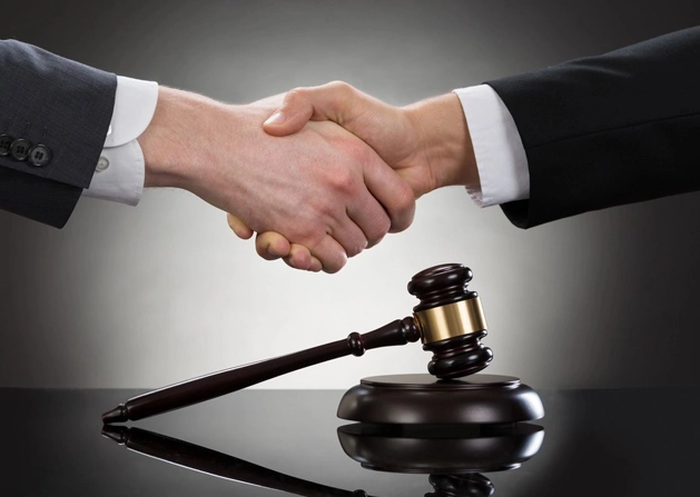 Two individuals in suits shaking hands over a gavel and sound block on a reflective black surface.