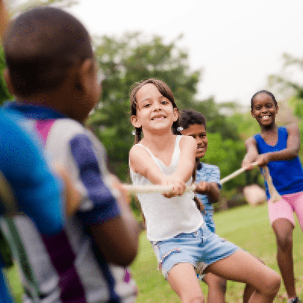 Children playing tug-of-war in a grassy park, smiling and pulling on a rope with trees in the background.