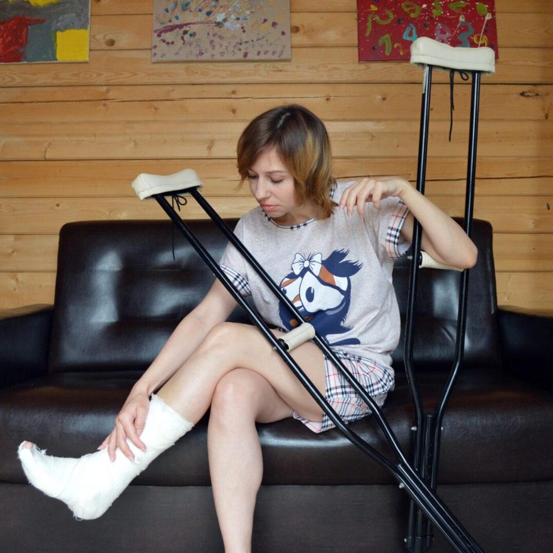 A person sits on a dark brown sofa with one leg in a cast and using forearm crutches. The background includes a wooden wall and several paintings.