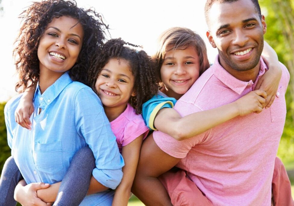A family of four smiles at the camera, with two children on the parents' backs in an outdoor setting.