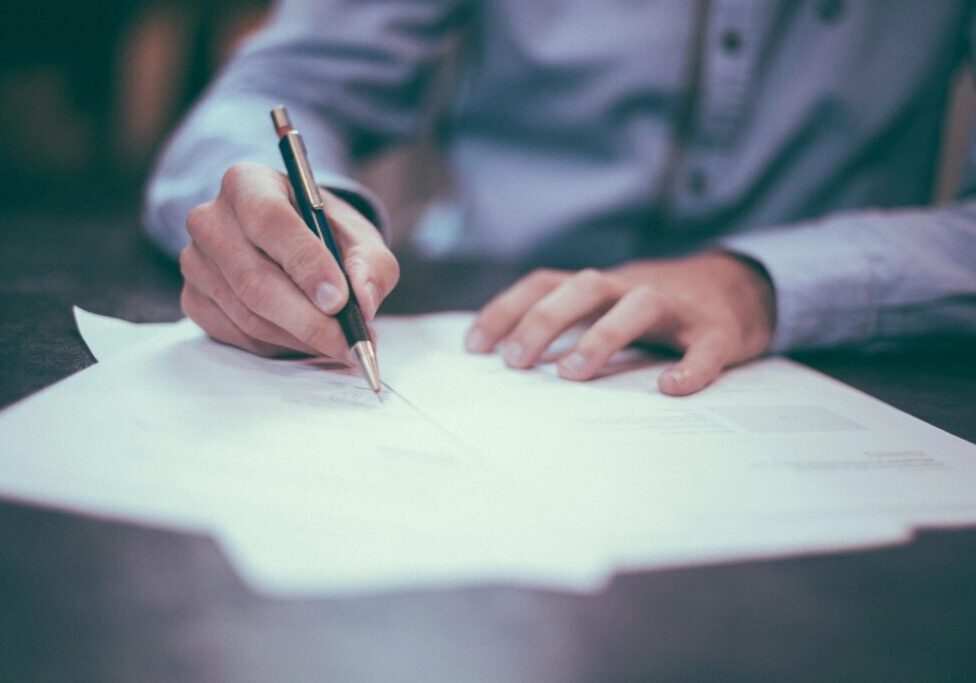 A person in a long-sleeve shirt is writing on papers with a pen while sitting at a desk.