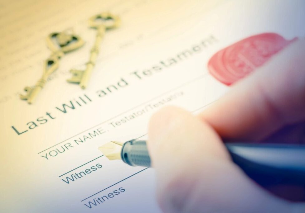 Close-up of a hand holding a pen, signing a "Last Will and Testament" document. There are two blank witness signature lines and a set of old-fashioned keys in the background.
