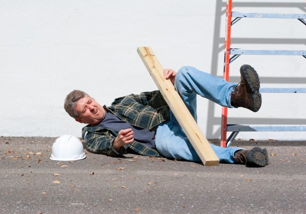 A man wearing a plaid shirt and jeans falls to the ground while holding a wooden board next to a ladder and a white hard hat.