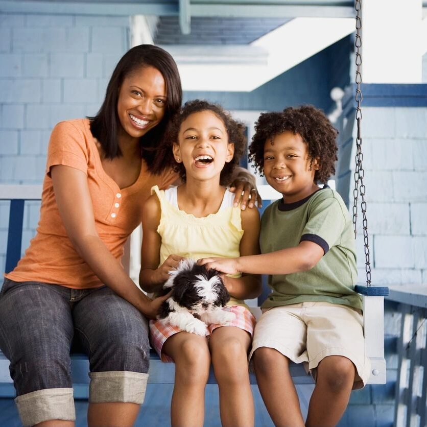 A woman and two children sit on a porch swing, smiling and holding a small black and white dog.