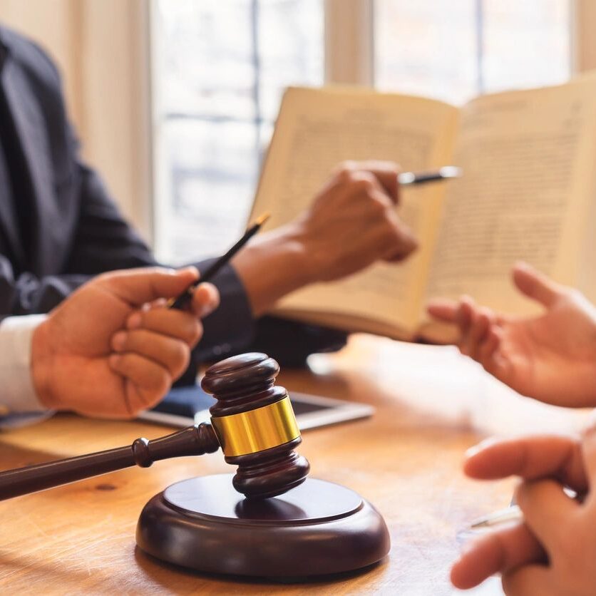 A gavel on a table with three people discussing and pointing at an open book in a room with large windows.