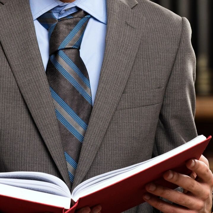 Man in a gray suit, blue shirt, and striped tie reading an open red book in a library setting with blurred bookshelves in the background.