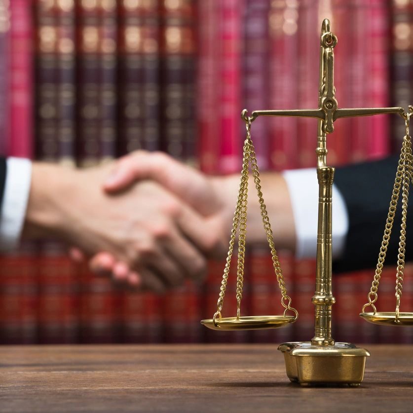 Two individuals shaking hands with a balance scale in the foreground and bookshelves in the background.