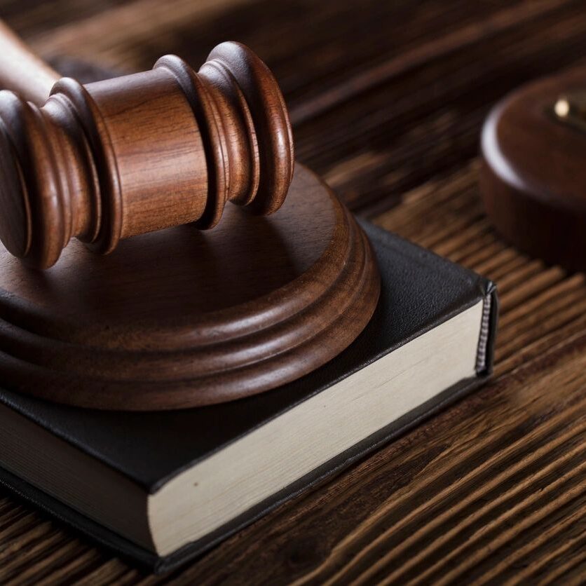 A wooden gavel rests on a closed book on a wooden surface, with a balance scale standing nearby.